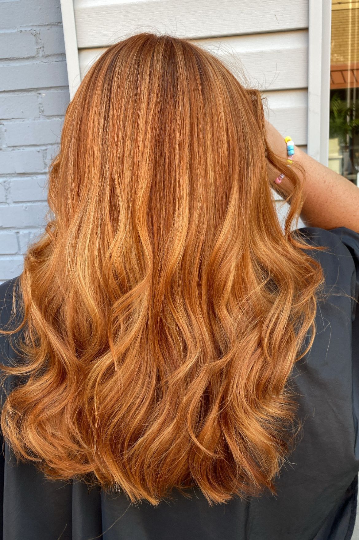 Woman with long, reddish-brown crimped hair, styled in waves.  Standing outdoors in front of a light blue wall.