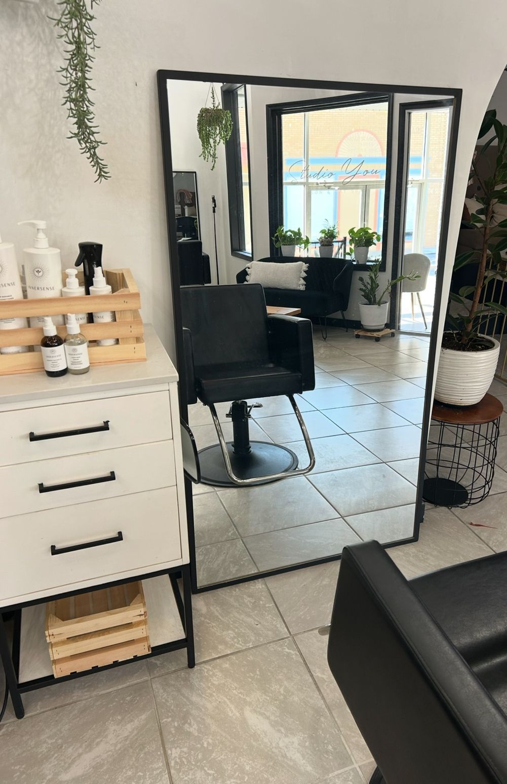 Hair salon interior with a large mirror reflecting a black chair, white walls, and a window.