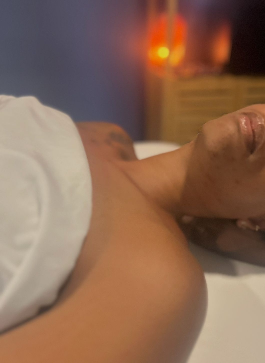 Person lying on a massage table, head to the side, wearing a white towel, and an orange light in the background.