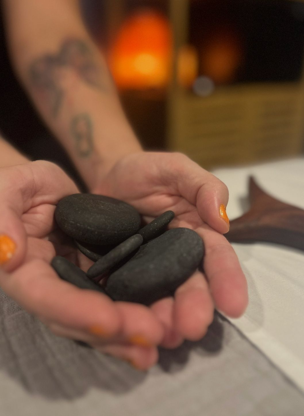 Person holding smooth, black stones, likely for massage, with orange nail polish. Background: warm lighting.