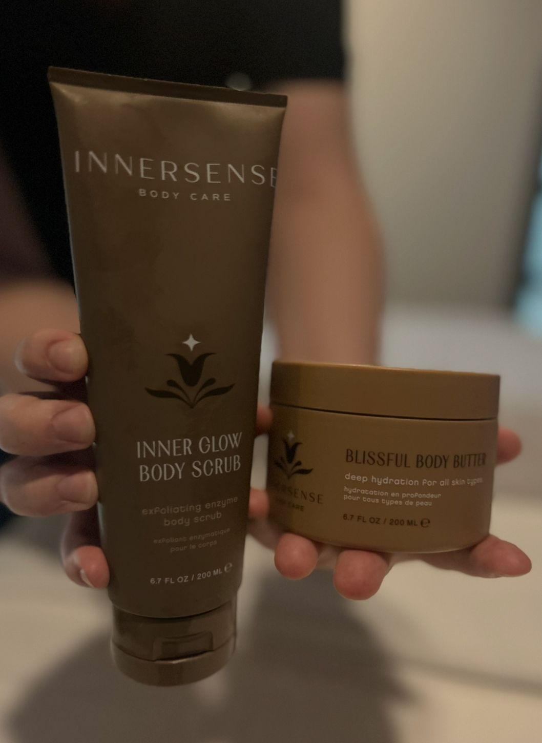 Person holding two brown Innersense body care products: body scrub and body butter.