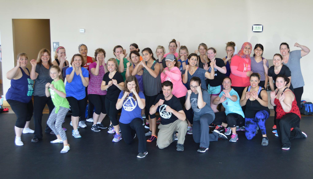 Group of people in workout attire, inside a gym, posing with fists up, smiling.