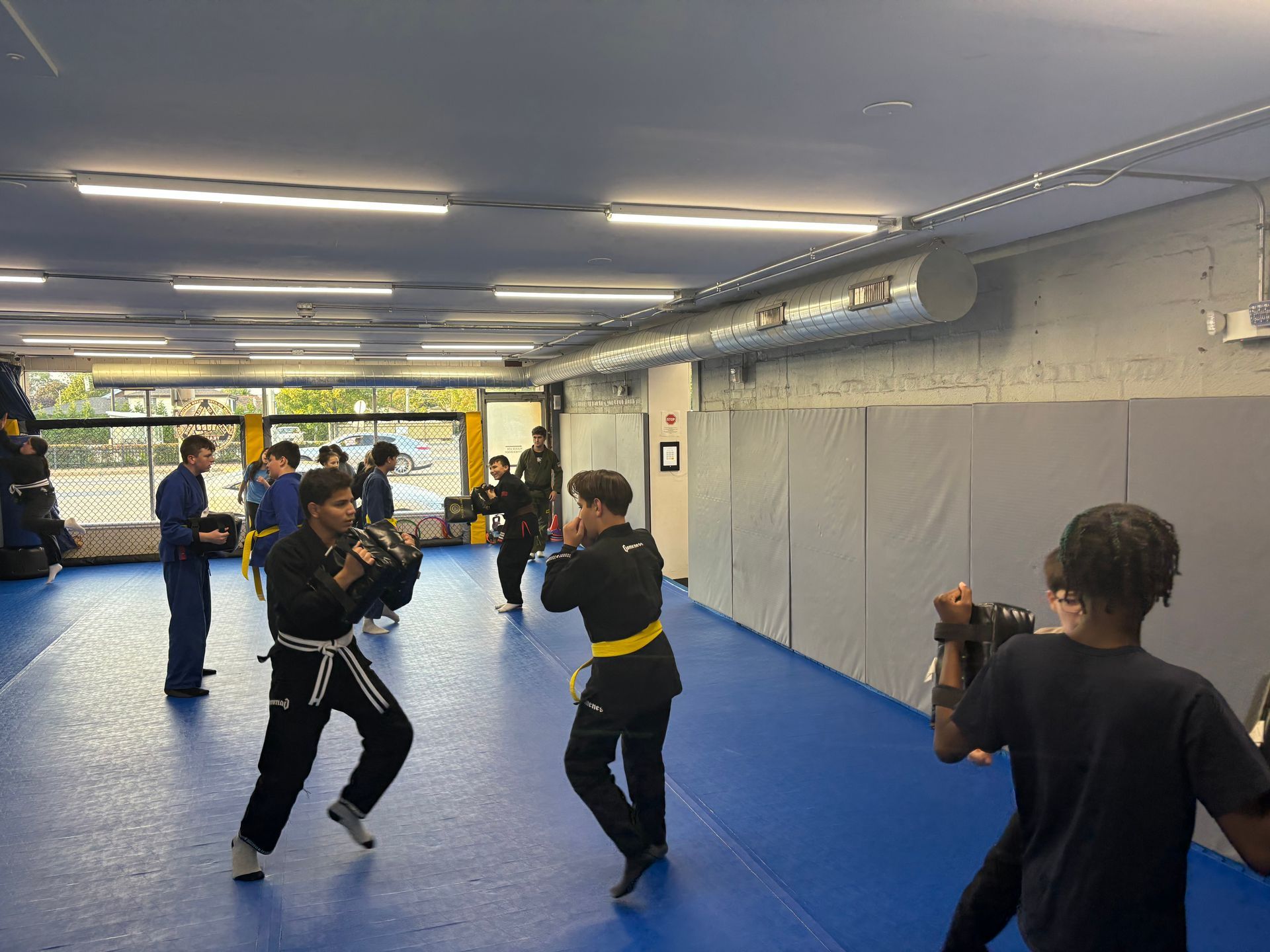 Martial arts training: People practicing kicks and punches in a gym with blue mats, wearing uniforms.