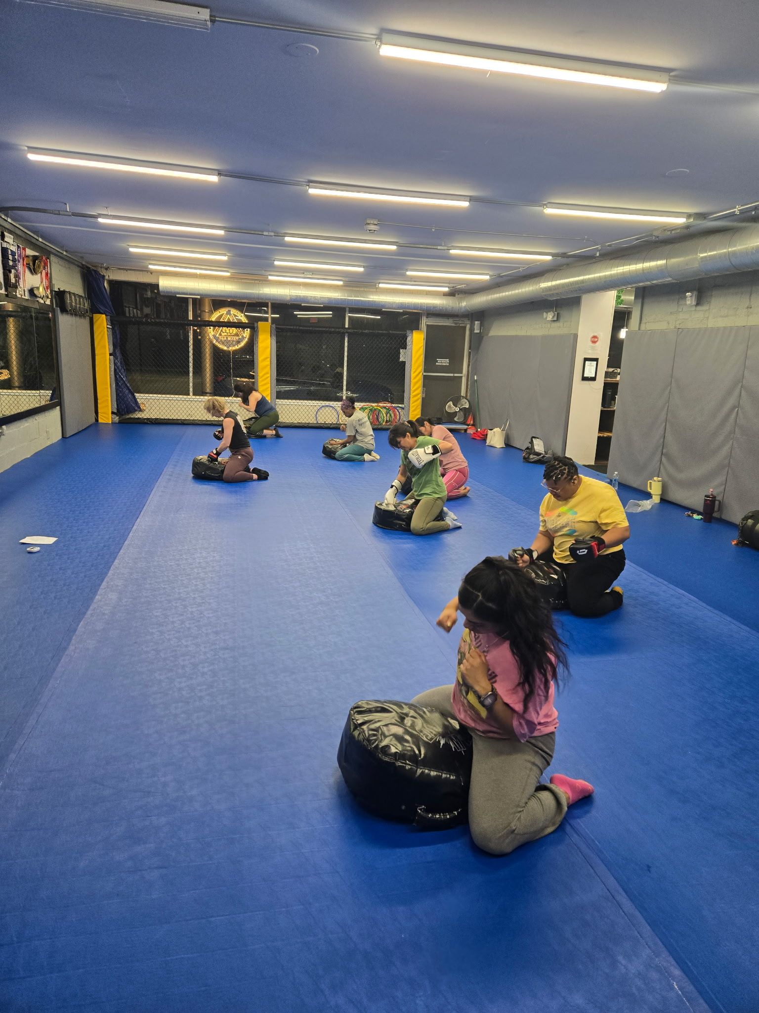 People in a martial arts class kneeling on mats. Blue mats, fluorescent lights.