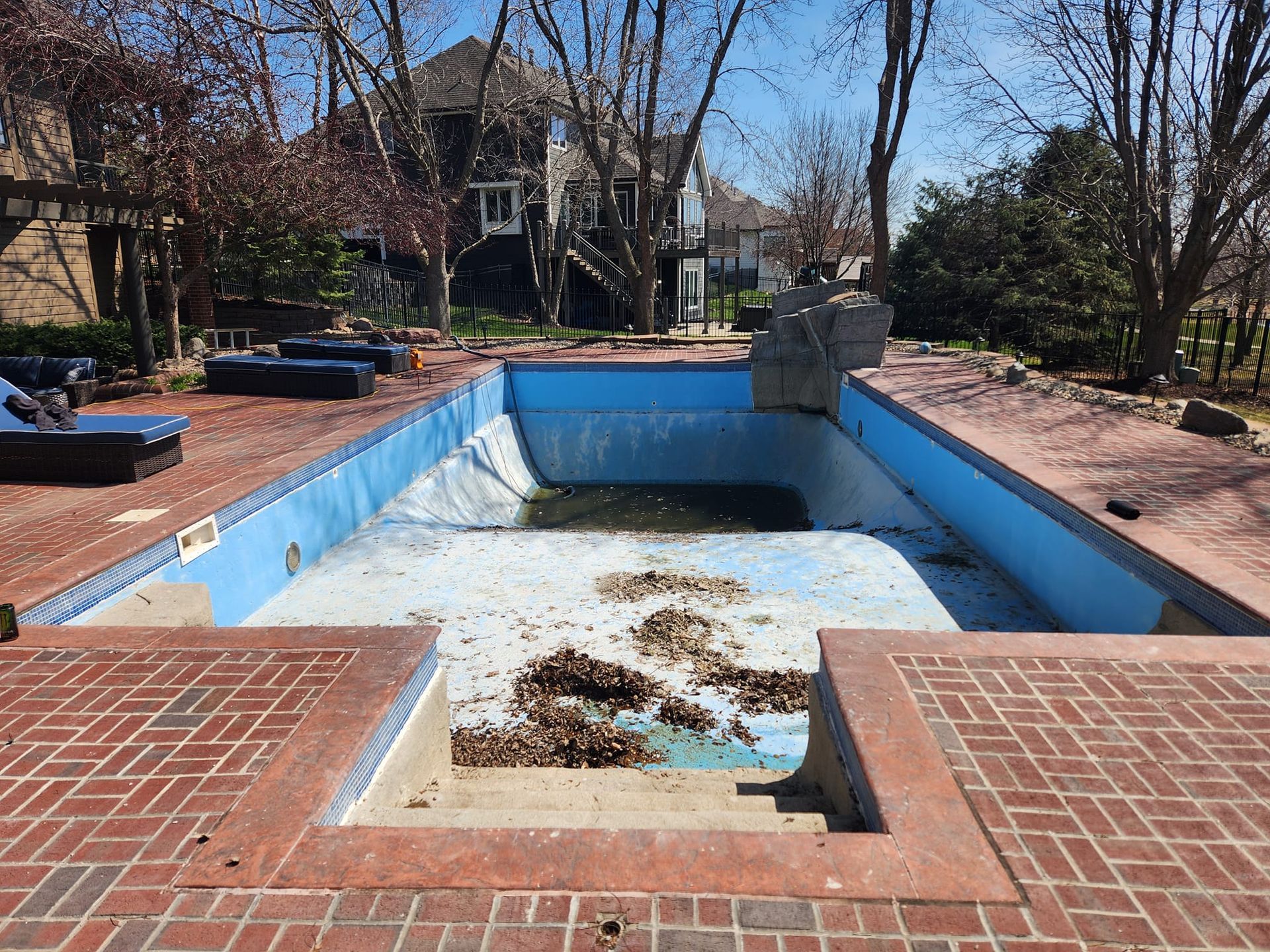 An empty swimming pool with a brick patio in front of a house.