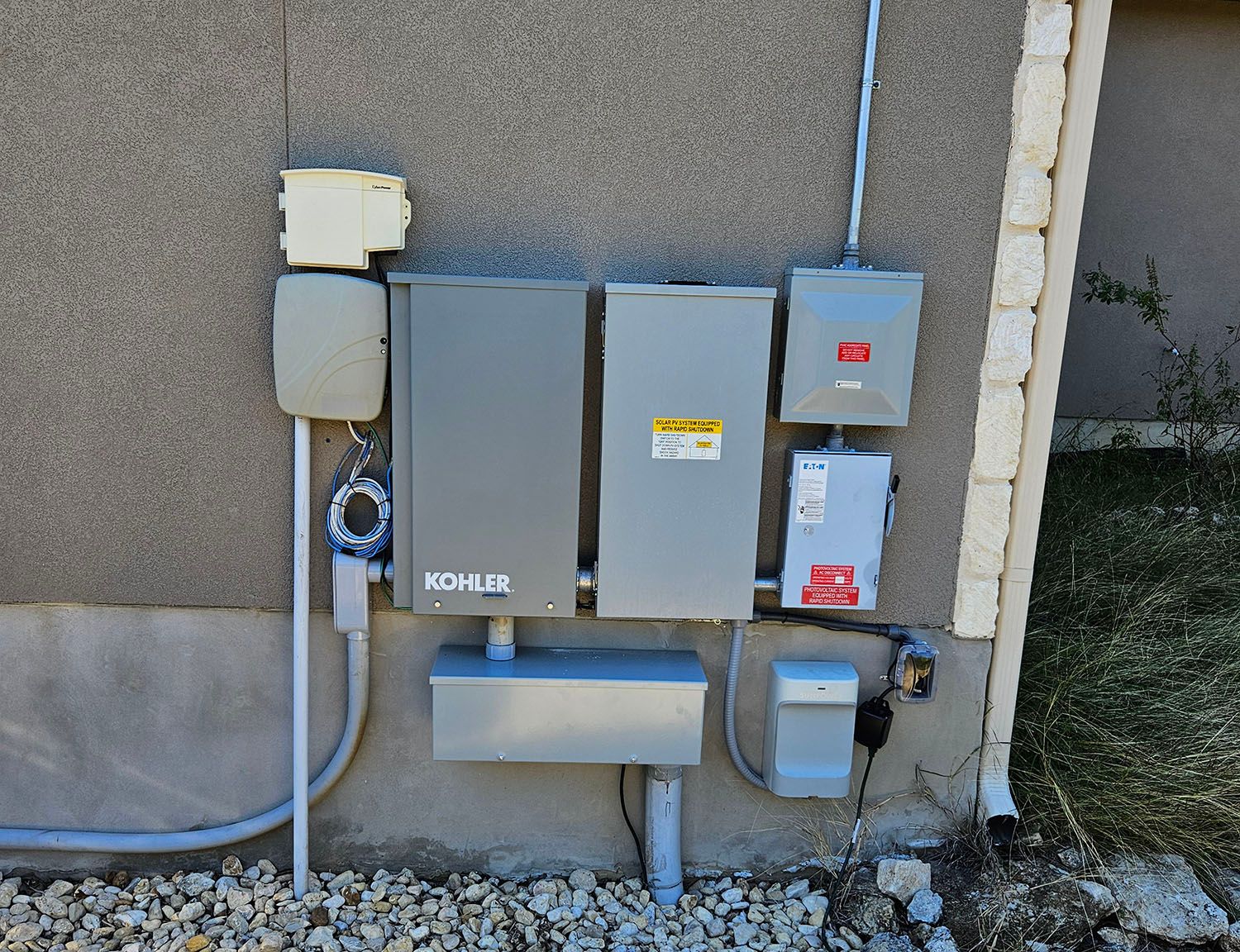 Outdoor utility meter and electrical boxes mounted on a wall beside conduit and gravel.