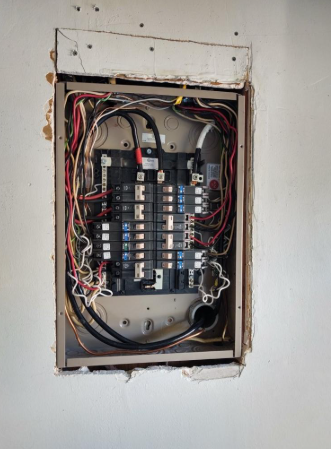 Open electrical breaker panel in a wall, with exposed wiring and circuit breakers.