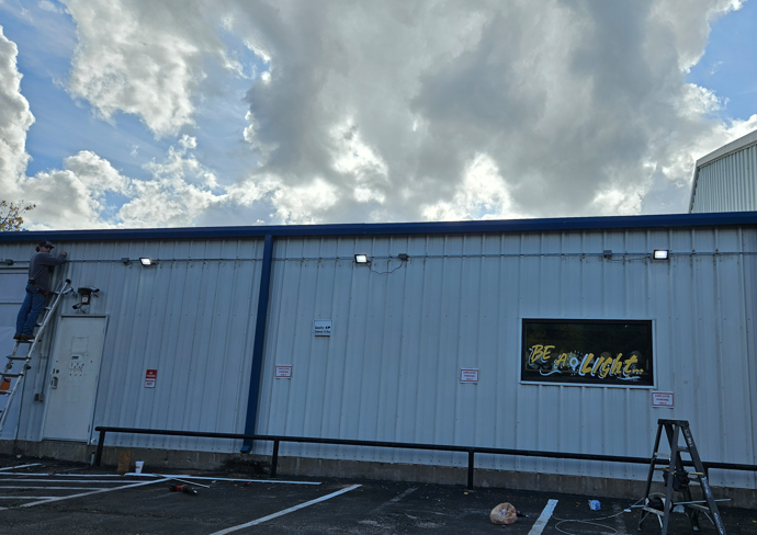 Industrial building with blue trim under cloudy sky, with a sign above a parking lot and ladder at right.