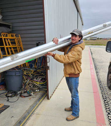 Man holding long silver metal pipes outside a workshop doorway