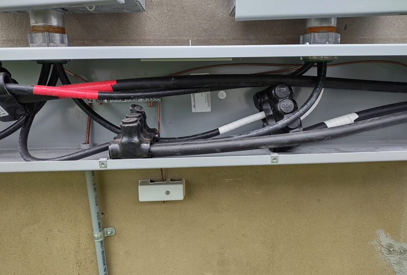 Open electrical panel with wiring, cables, and red conduit under a box outside a beige wall