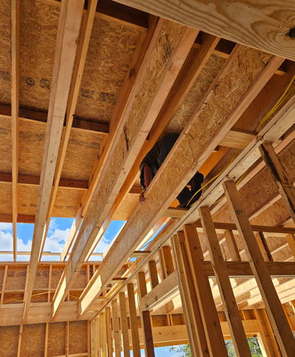 Wood-framed house under construction, with exposed roof trusses and plywood sheathing.