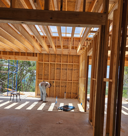 Wood-framed house under construction with exposed studs and a worker bending inside.