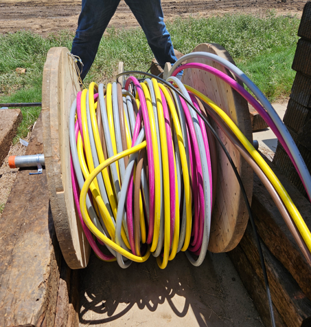 Coiled pink, yellow, and gray hoses on a large wooden spool outdoors on bare ground