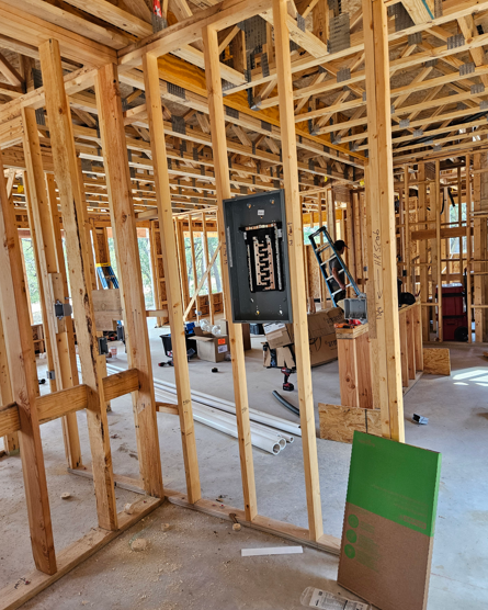 Interior of a house under construction, showing exposed wood framing, wiring, and a panel in progress.