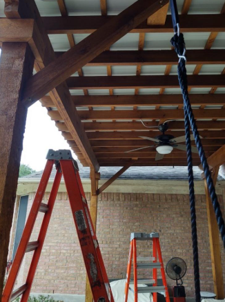 Covered patio with two red ladders, wooden beams, a ceiling fan, and hanging chain work under construction