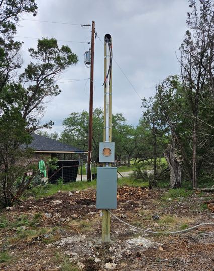 Outdoor utility pole with a blue electrical box in a grassy yard, with trees and a house in the background