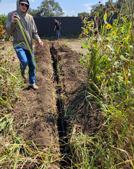 Person in a sun hat stands beside a freshly dug trench in an overgrown field, holding a long tool.