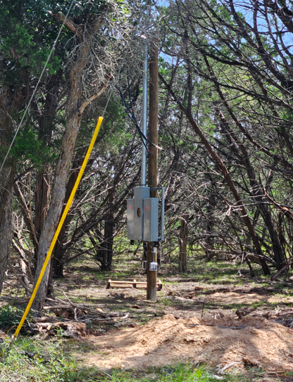 Utility pole with electrical box in a wooded area, marked by a yellow pole beside it.