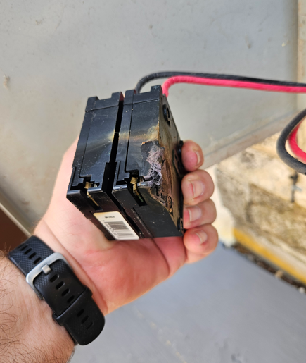 Hand holding a black battery pack with red and black wires attached, near a wall-mounted device