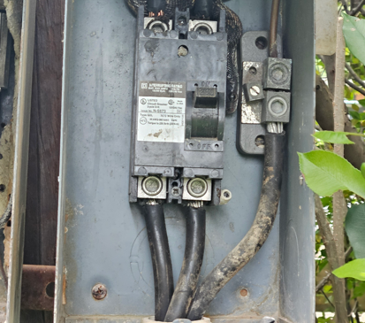Open electrical breaker panel with thick black cables and weathered wiring outdoors