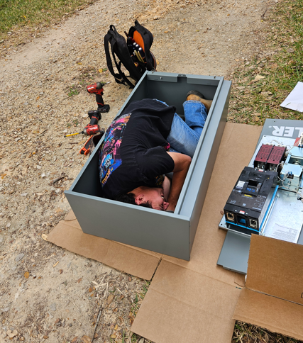 Person lying in a gray cardboard box outdoors beside tools and electronics on cardboard sheets.