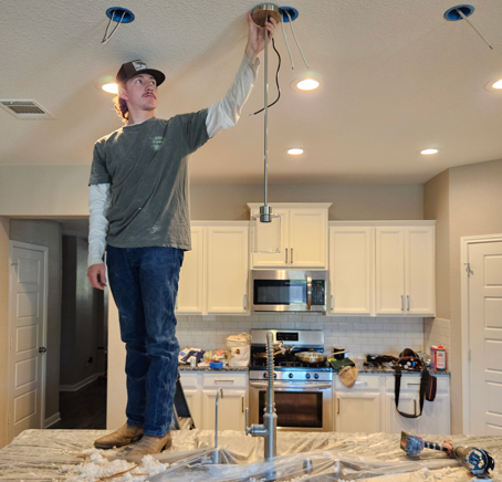 Man hanging a ceiling light in a kitchen with cabinets and appliances, standing on a covered countertop.