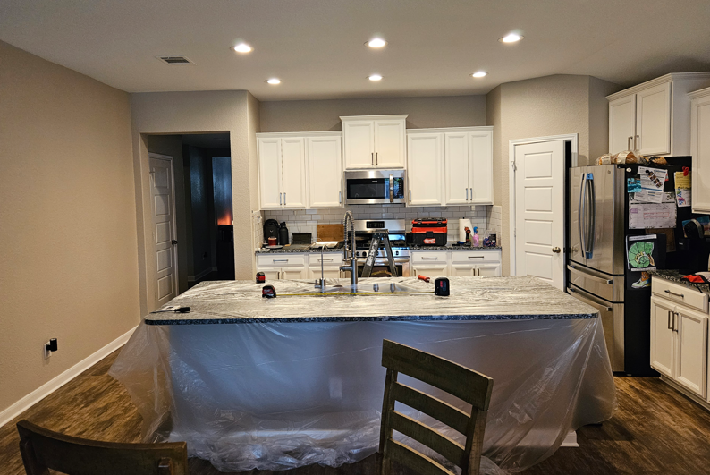 Kitchen under renovation with white cabinets, granite island wrapped in plastic, and appliances along the back wall
