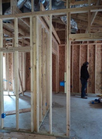 Interior framing of a house under construction, with exposed wood studs and one worker standing near the back wall.