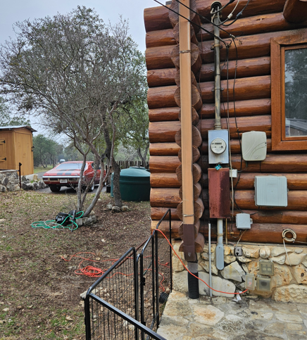 Log cabin exterior with utility meters, a dirt yard, trees, and a parked car in the background