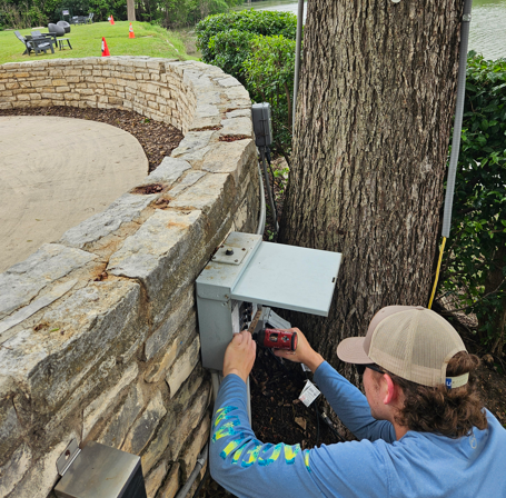 Technician crouches by a stone wall, working on a gray utility box near a tree outdoors.