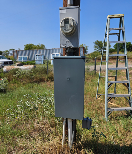 Gray electrical meter box on a post in a grassy field, with a ladder standing beside it.