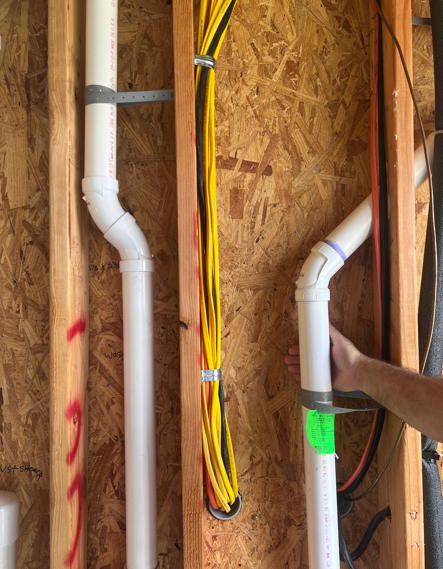 Two white PVC pipes in a wall cavity with yellow electrical wiring and a hand pointing at the right pipe.