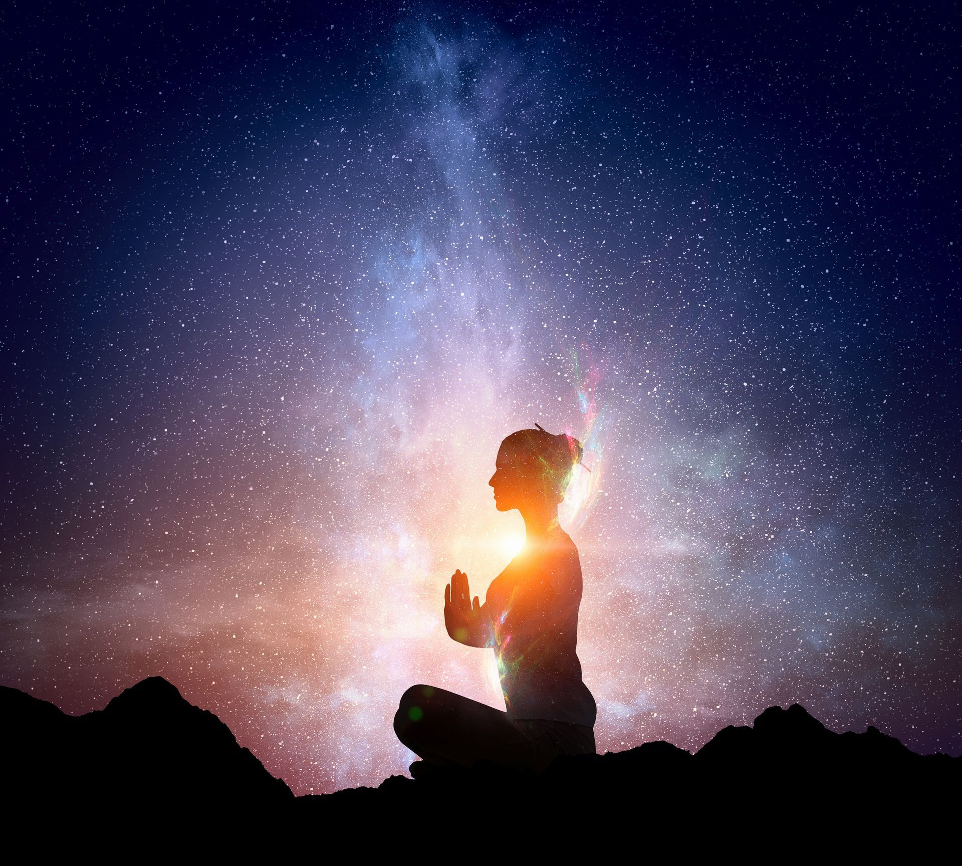 Silhouette of person meditating in lotus position, hands together, under starry night sky with glowing light.
