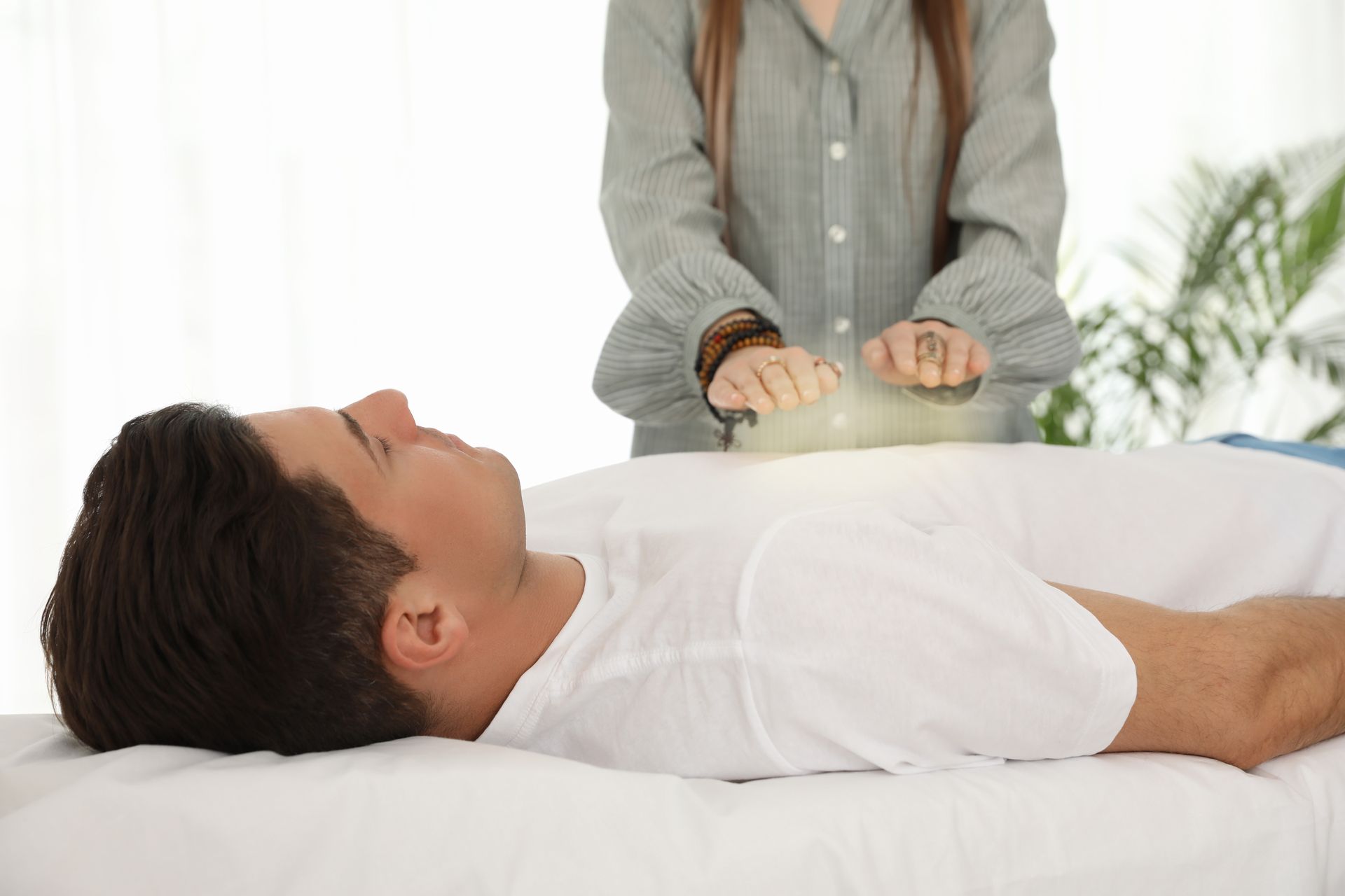 Man receiving reiki treatment; practitioner's hands above his abdomen. White shirt and sheet, bright room.