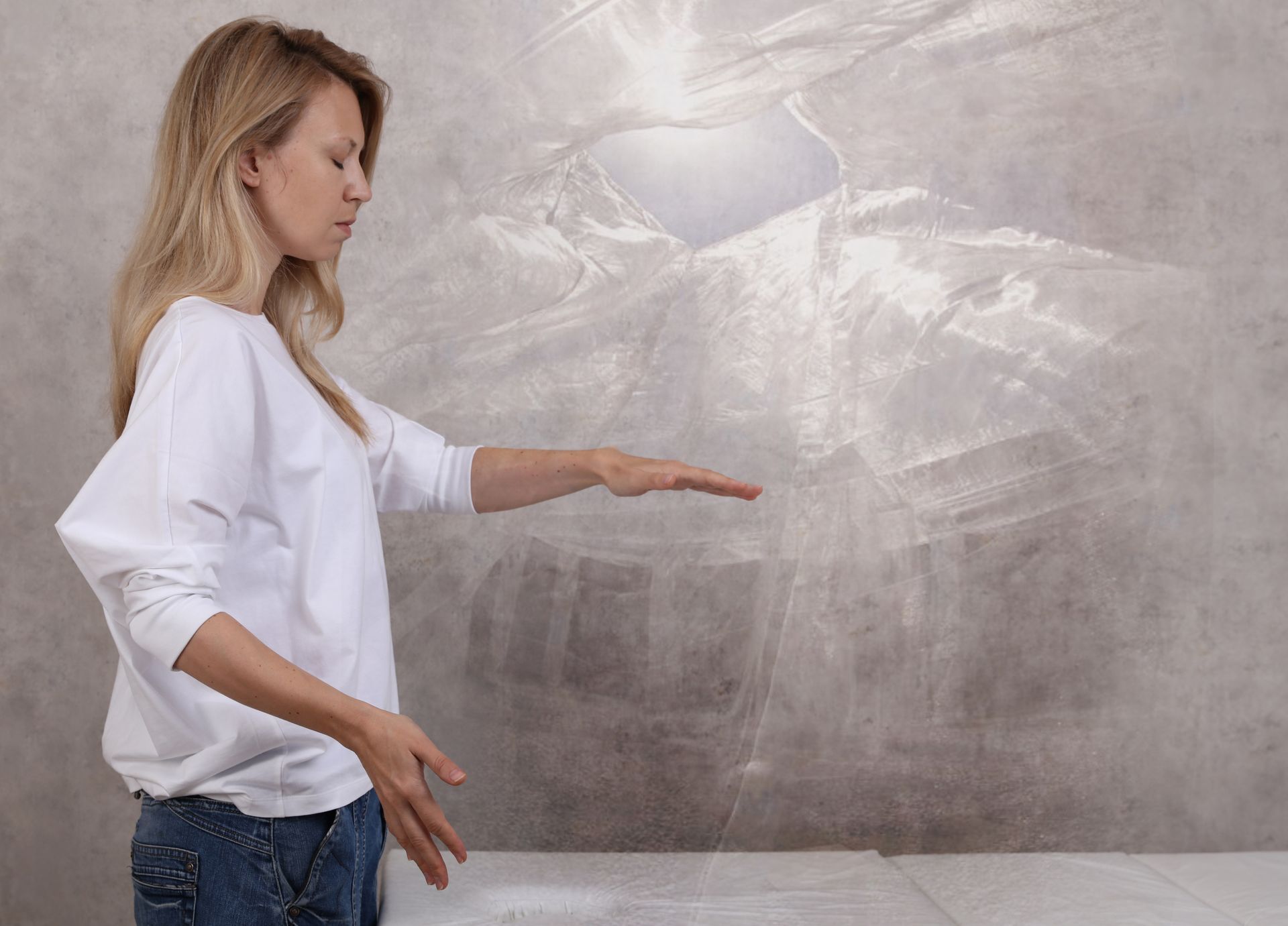 Woman with blonde hair, arms outstretched, meditating against a textured gray wall, white light above.