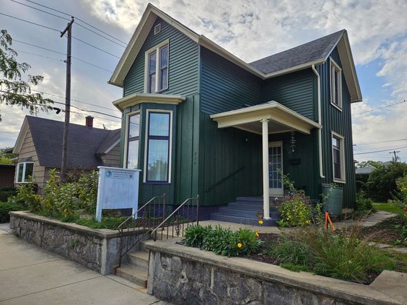 Green two-story house with a porch and a white sign in front of a garden.