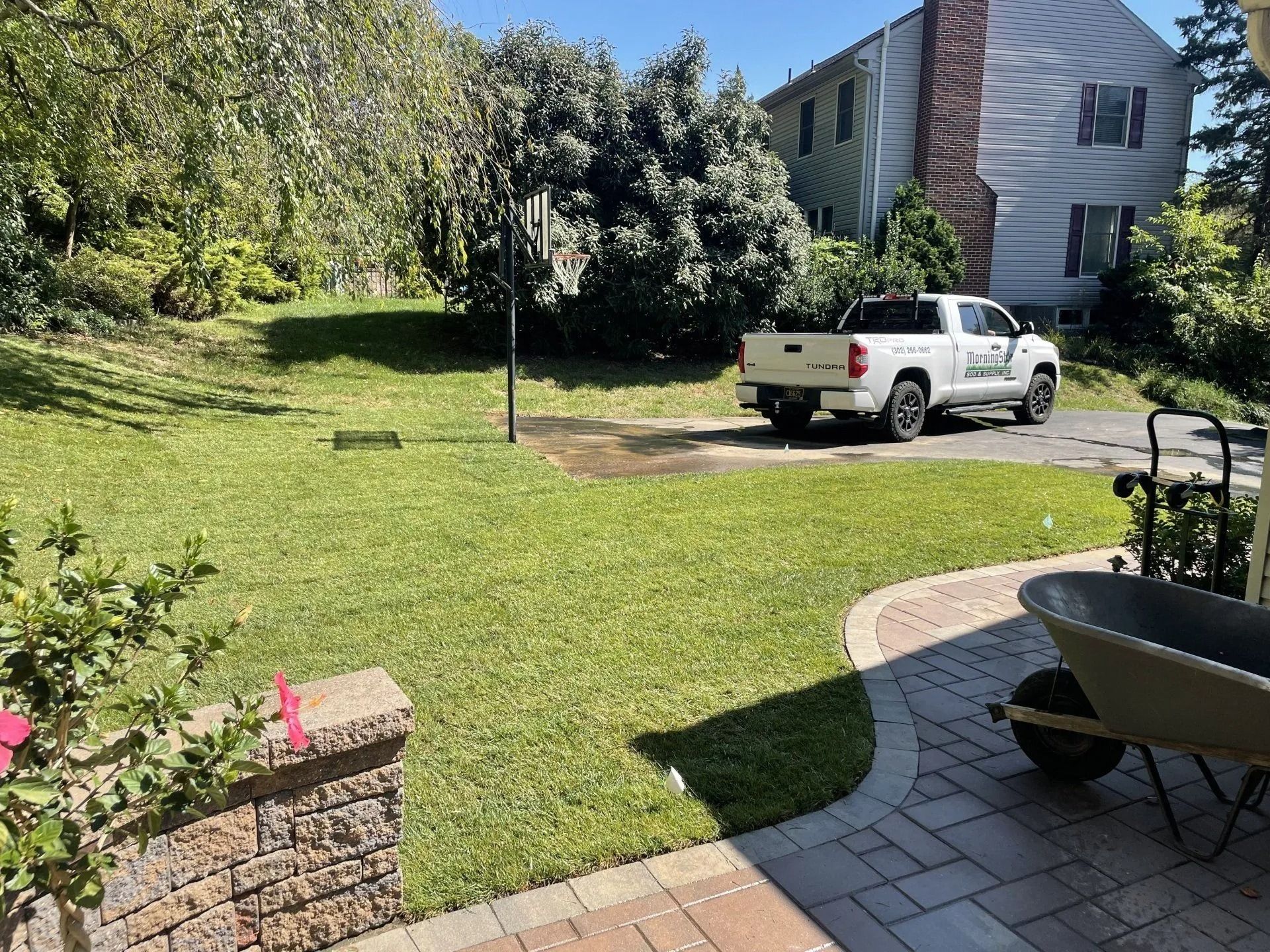 A white pickup truck parked in a grassy yard next to a brick-paved patio with a wheelbarrow.