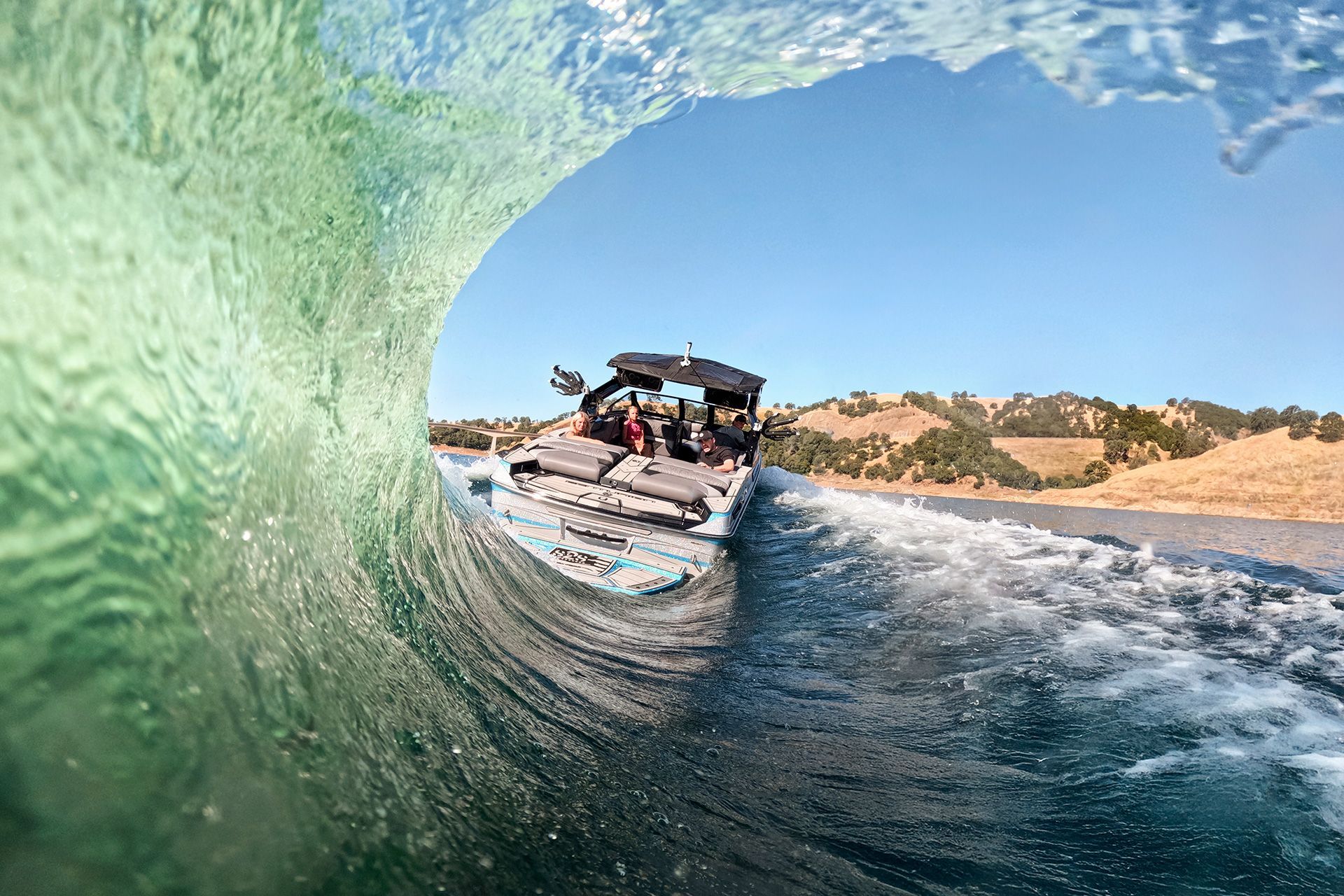 View from inside a breaking wave, looking out at a boat on a lake; clear water, blue sky, and brown hills.