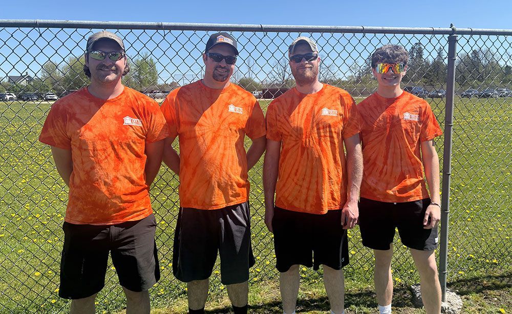 Four men in orange shirts are standing next to each other in front of a chain link fence.