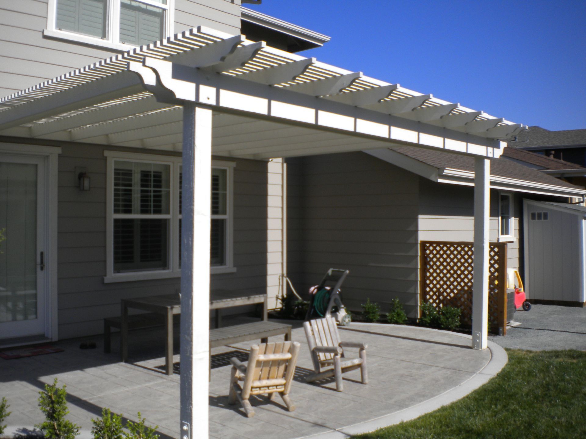 Patio with white pergola, chairs, and grill beside a house and lawn