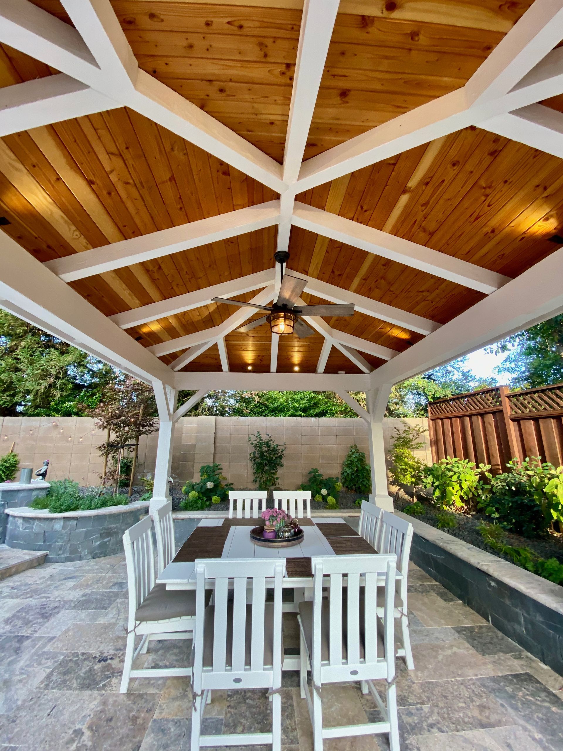 Covered patio dining area with white table and chairs, wooden ceiling, and ceiling fan in a landscaped backyard