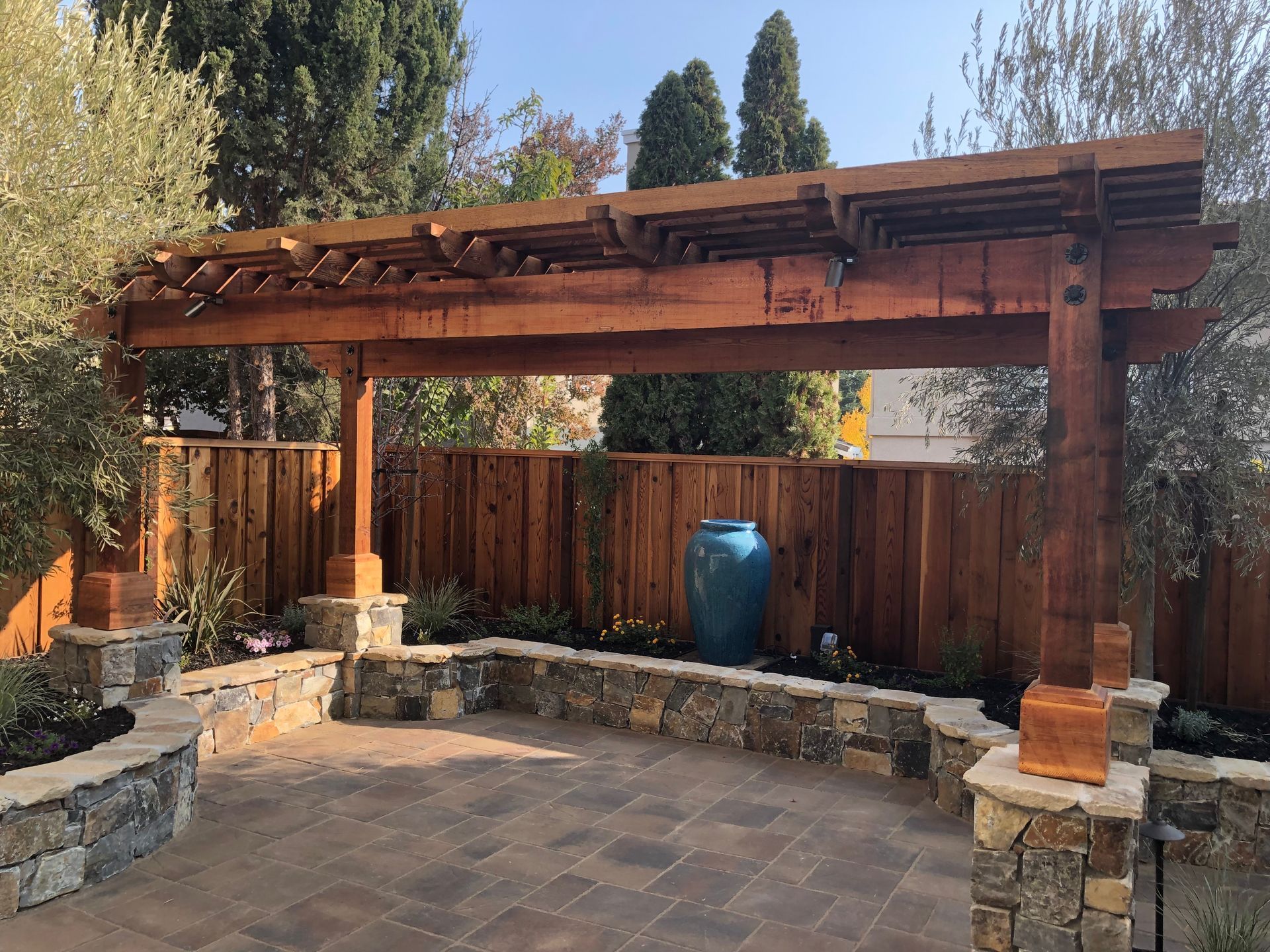 Wooden pergola over a stone patio with built-in seating and a fenced garden backdrop