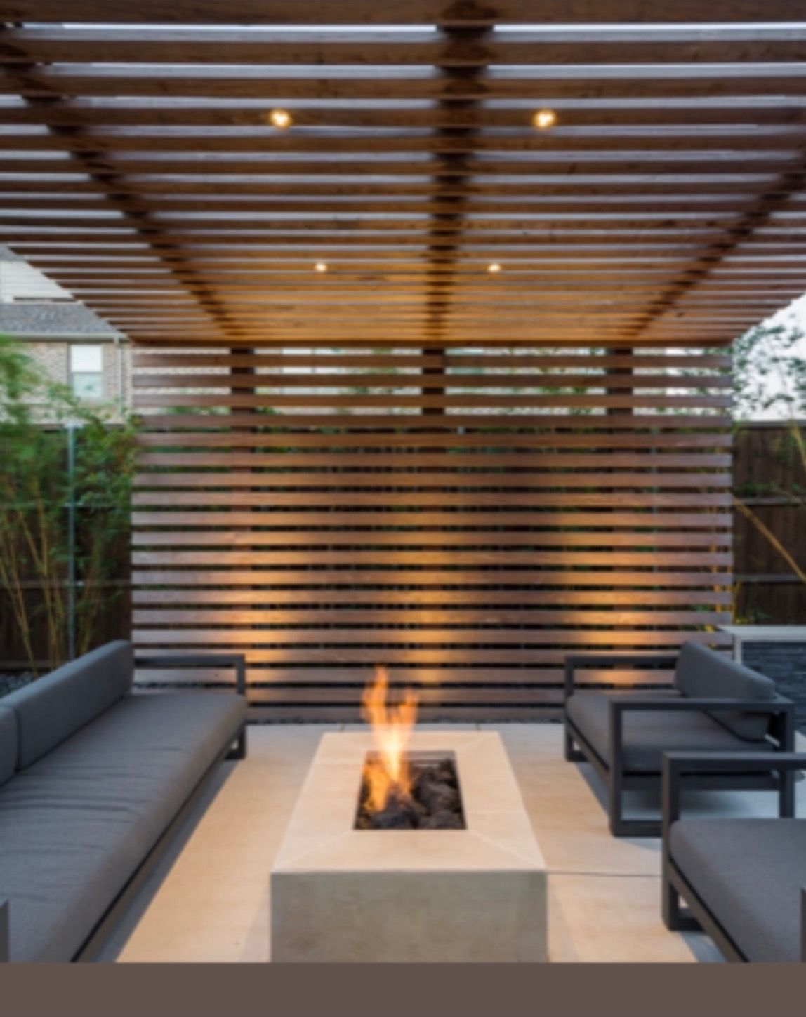 Modern outdoor patio with gray sofas, a fire pit, and a wood slat pergola overhead.