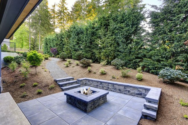 A stone patio featuring a square fire pit, a low retaining wall, and steps leading to a gravel path in a landscaped yard.