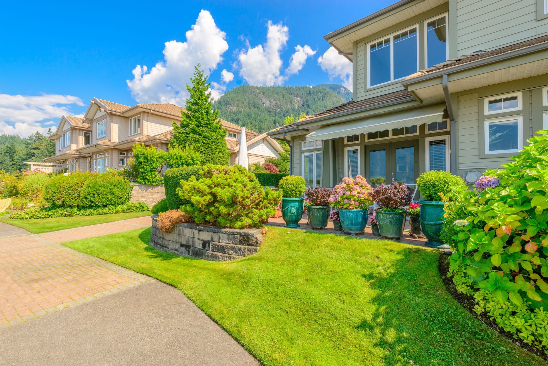 A suburban home with a green lawn, potted plants, and a backdrop of mountains under a blue sky.