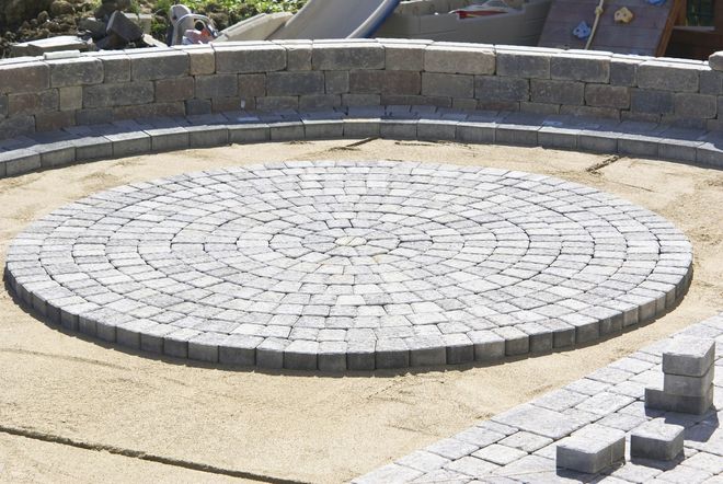 A circular stone paver patio under construction, surrounded by a low stone retaining wall on a sandy foundation.