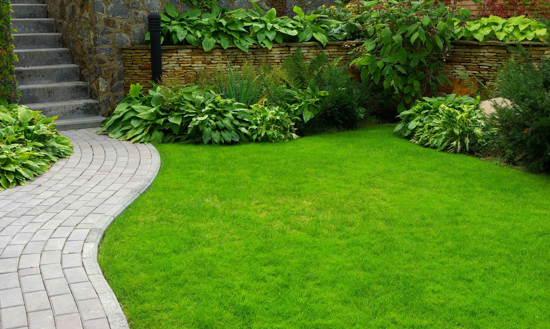 A curved stone path leads to a stone staircase next to a lush green lawn and landscaped garden beds.