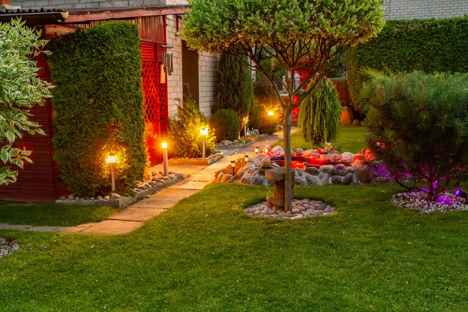 A stone path through a lush garden illuminated by warm yellow ground lights and small purple accent lights at twilight.