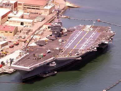 Aircraft carrier docked at a pier; gray ship, black runway markings, buildings, harbor.