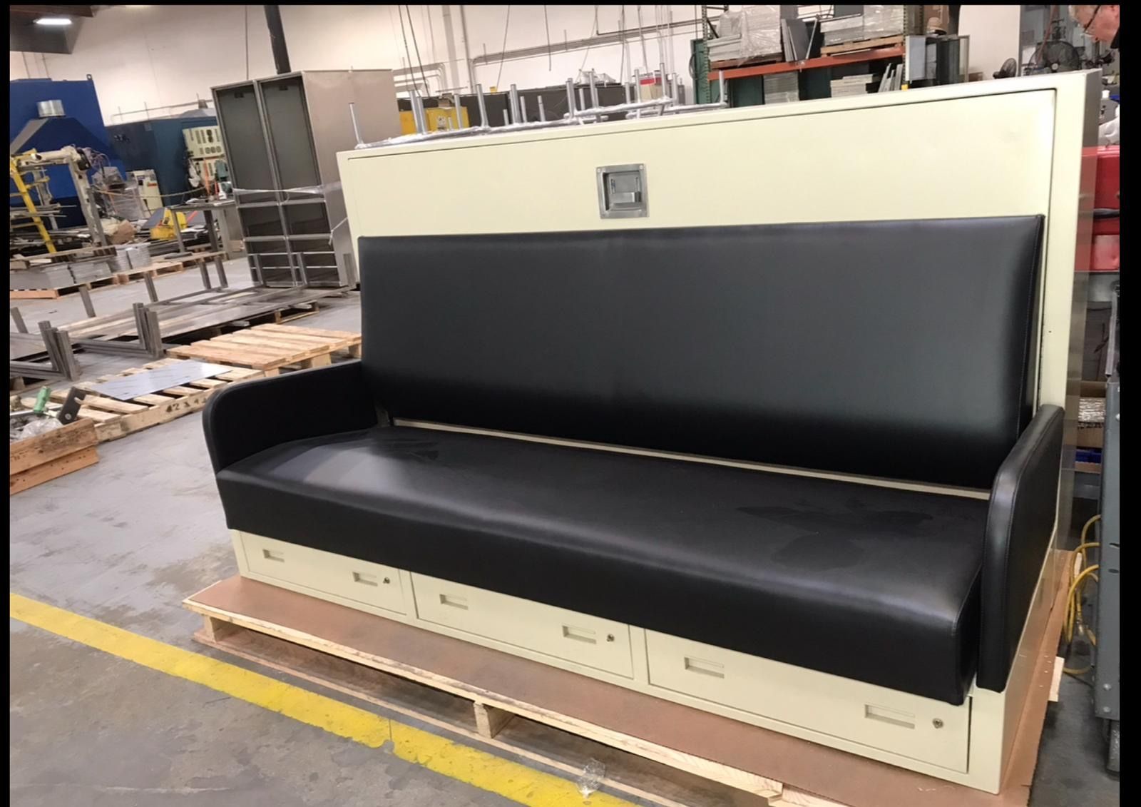 A beige and black Murphy bed/sofa combo with drawers, set in a manufacturing environment.